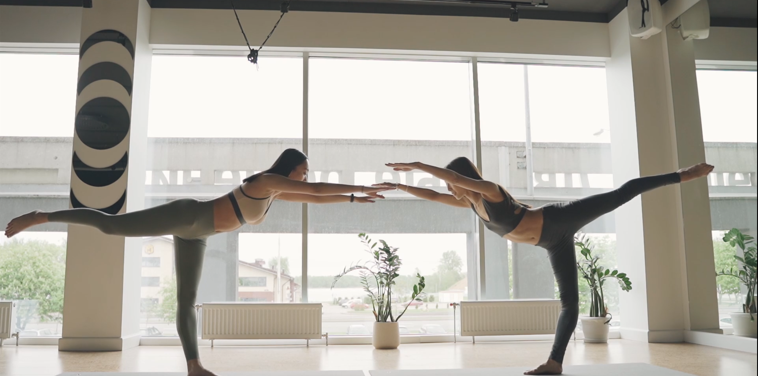 Two women practicing Warrior III Yoga Pose (Virabhadrasana III) in a bright studio with large windows, demonstrating balance, strength, and focus