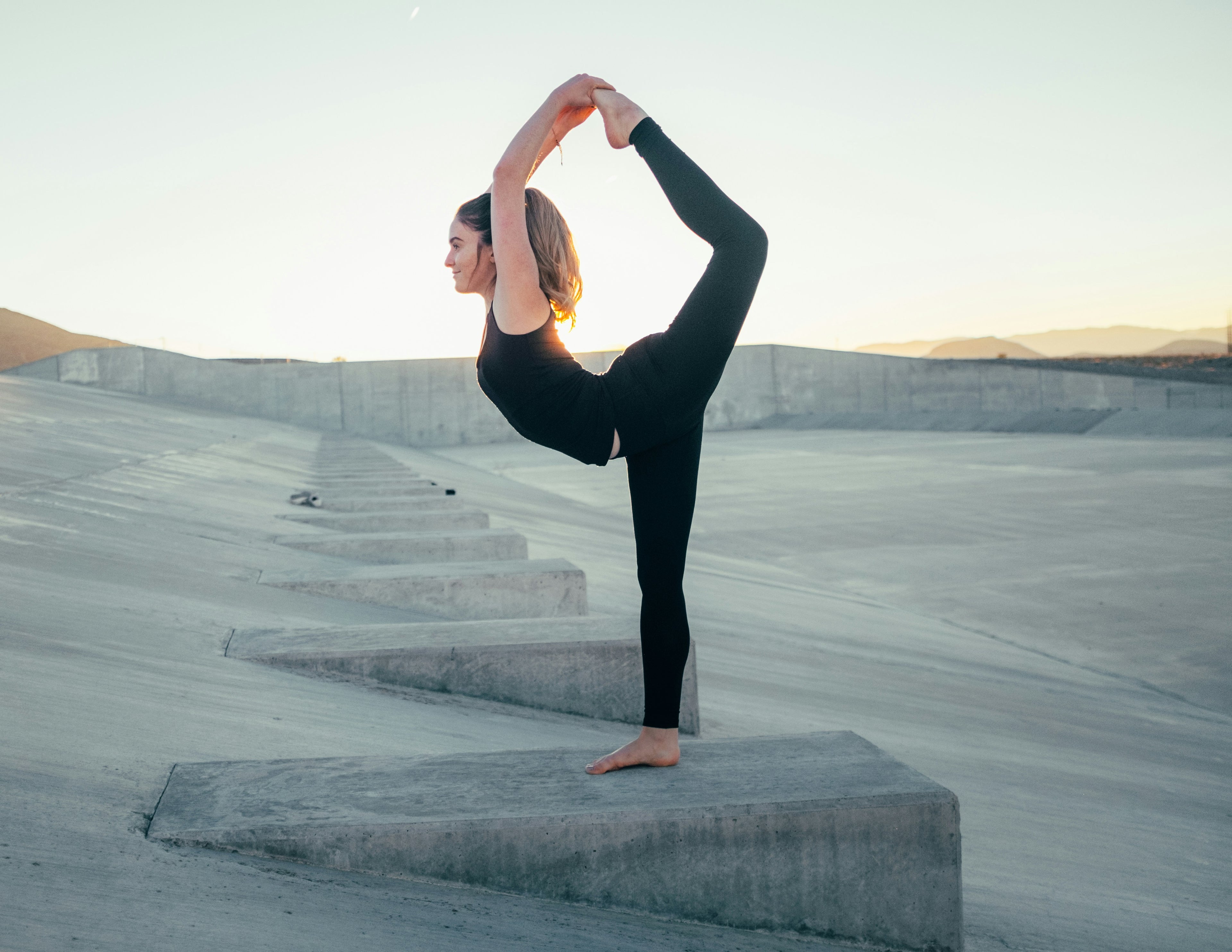 A woman practicing yoga outdoors in a flexibility pose at sunset.