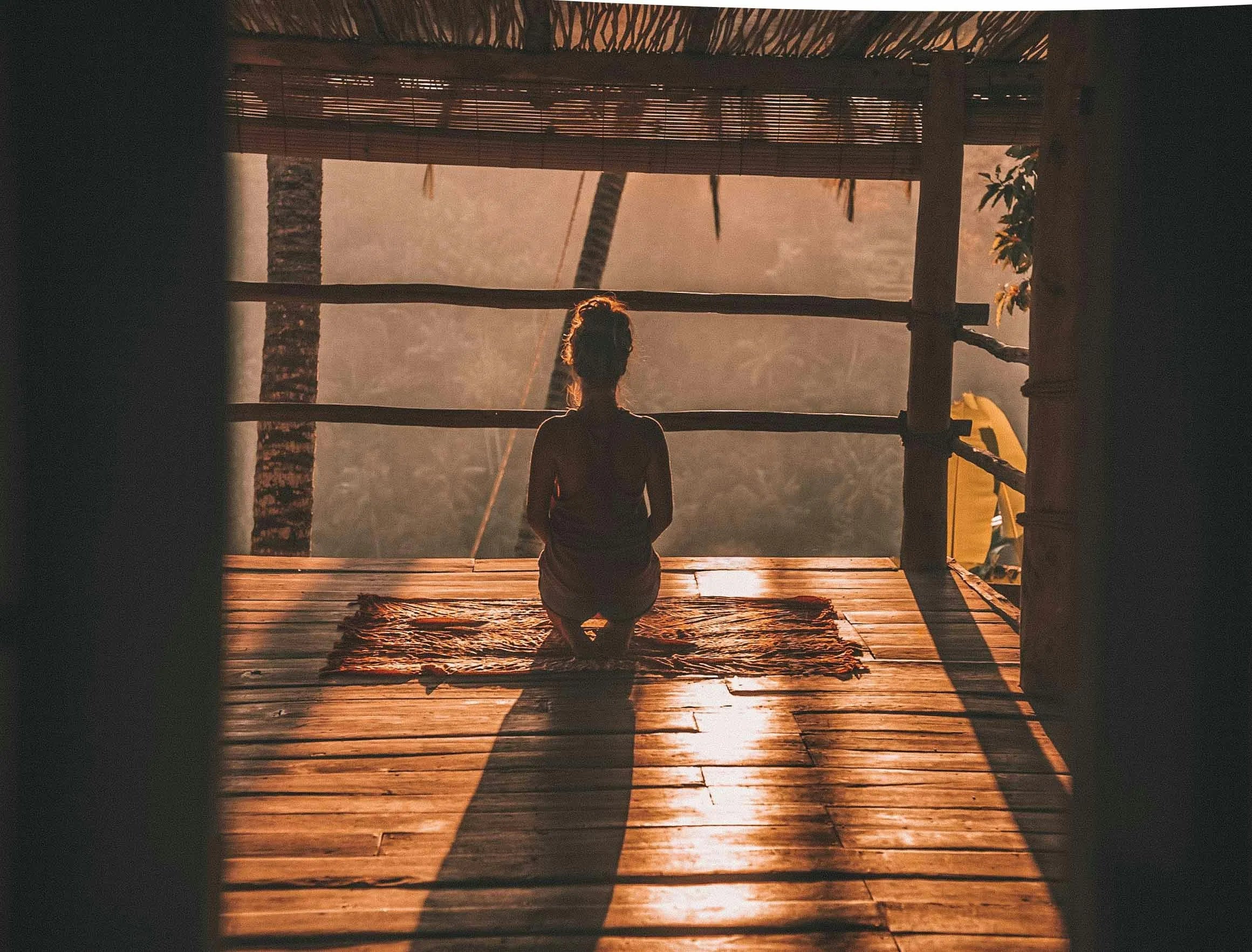 A girl sitting in the middle of a grass house in the sunset, her back facing us